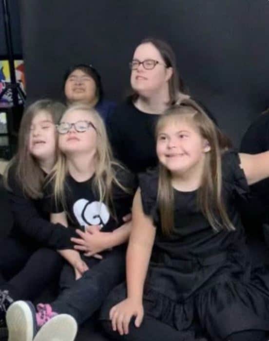 A group of five children and one adult, all with Down syndrome, sit together in front of a dark backdrop, smiling and posing for a charity photo. Everyone is wearing black clothing.