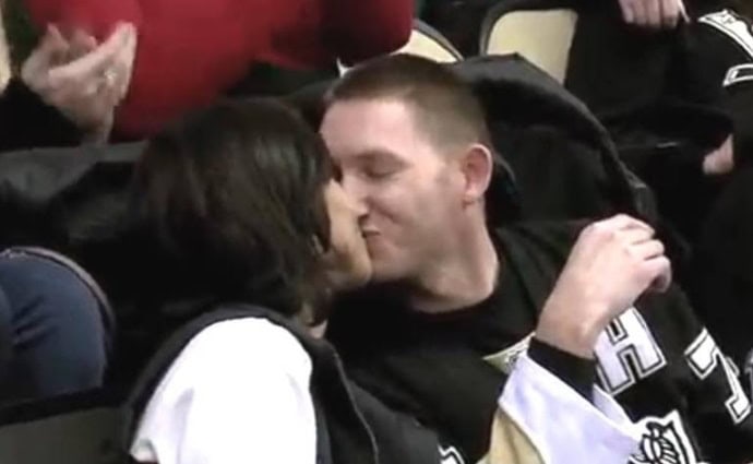 A couple sits close together in a stadium, smiling and sharing a kiss. Both are wearing dark sports jerseys, and the spirit of giving is felt among other spectators visible in the background.