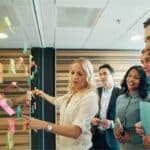 A group of five professionals stand by a glass wall covered with colorful sticky notes. A woman points to one note while the others listen attentively, suggesting a collaborative meeting focused on charity and giving.