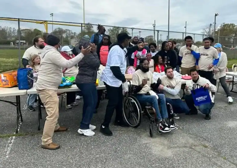 A diverse group of people, including a man in a wheelchair, gather outdoors around tables with food and supplies for a charity event. They are smiling, talking, and posing near a sports field with a fence and goalposts on a cloudy day.