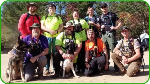 A group of ten people, some wearing bright shirts and outdoor gear, pose together outdoors with two dogs in front—gathered in the spirit of giving. The background features trees and sunlight.