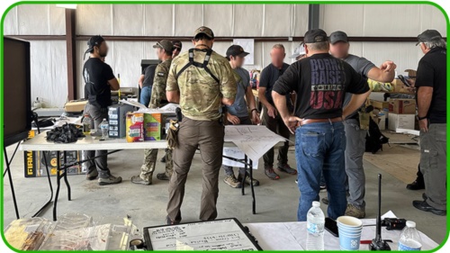 A group of people in tactical clothing gather around a table with maps and supplies in a warehouse-like setting, discussing plans to provide help. Tables hold gear, radios, water bottles, and boxes for charity. Faces are blurred for privacy.