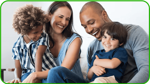 A happy family of four sits closely together, smiling and laughing. The parents sit in the middle, each holding a young child, creating a warm and joyful atmosphere filled with love and giving.