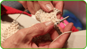 Close-up of hands holding knitting needles and working with cream-colored yarn, creating a textured knitted fabric—a gentle reminder of the warmth that giving and charity can bring. Green border frames the image.
