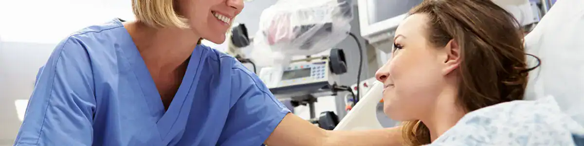 A nurse in blue scrubs smiles and talks to a patient lying in a hospital bed at the Center For Assault Treatment Services (CATs), creating a caring and supportive atmosphere focused on compassionate assault treatment.