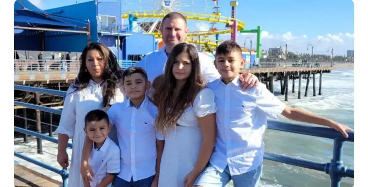 The Vandenbroucke Family of six stands together on a pier by the ocean, all dressed in white and light colors. A blue building and a colorful amusement park ride are visible in the background on this sunny day.