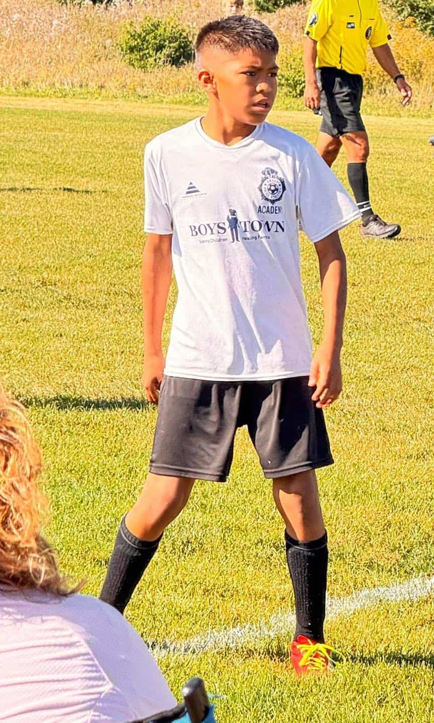 A young boy in a white soccer jersey and black shorts stands on a grassy field, looking to the side. At just 14, he dreams big—as does the volunteer in a yellow referee shirt seen in the background.