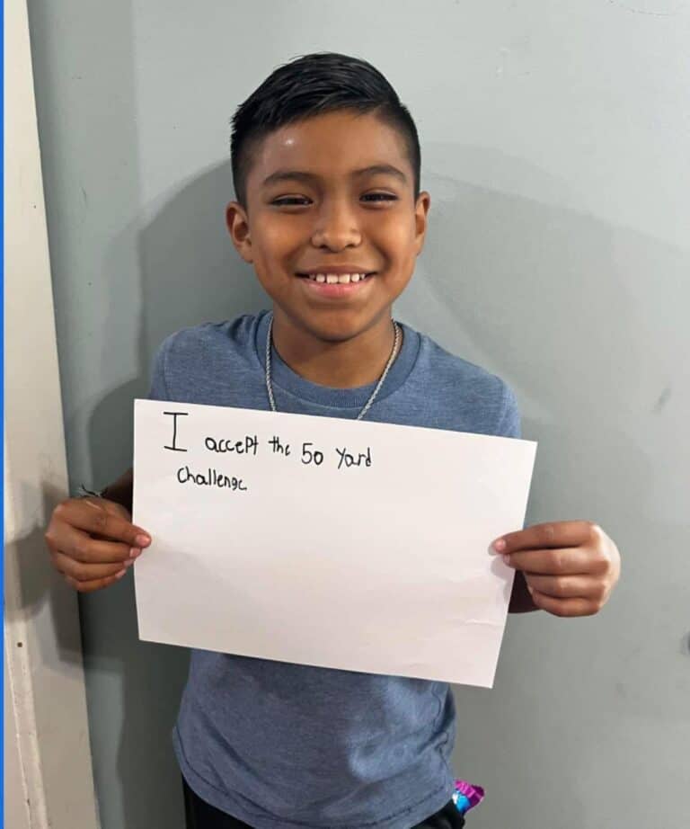 A smiling 14-year-old boy in a blue shirt holds a handwritten sign that says, I accept the 50 yard challenge, standing against a light gray wall, ready to volunteer.