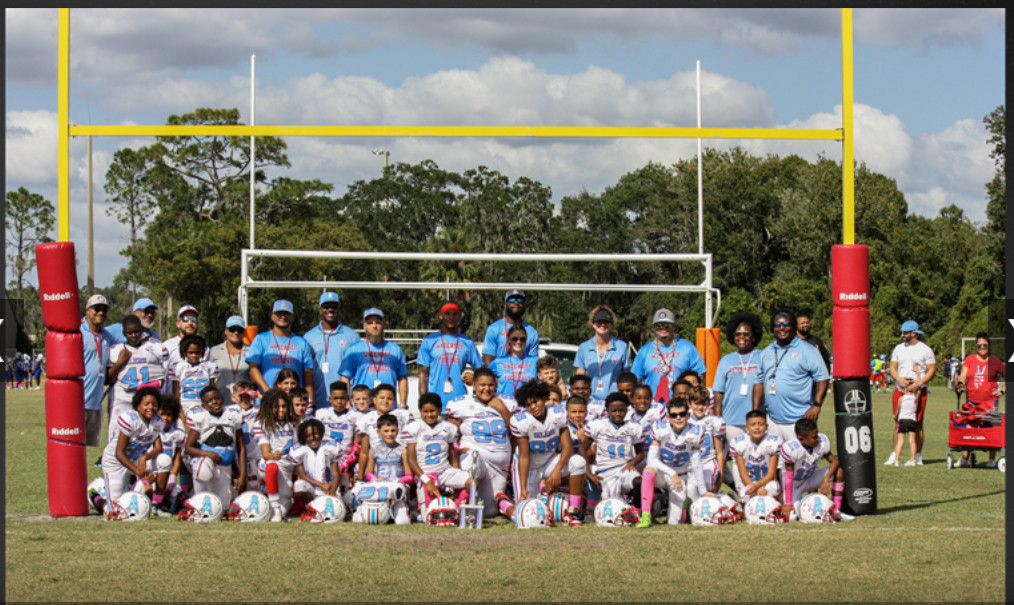 A Youth Sports football team and coaches pose together on a grassy field in front of a goalpost, all wearing Warterset Oilers uniforms and smiling for a group photo. Trees and several adults are visible in the background.